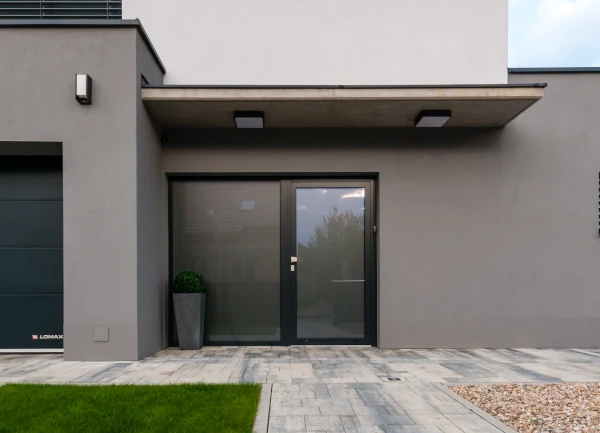 Moderne Haustür aus Glas und Aluminium in grauer Hausfassade mit Vordach und Pflastersteinen.
